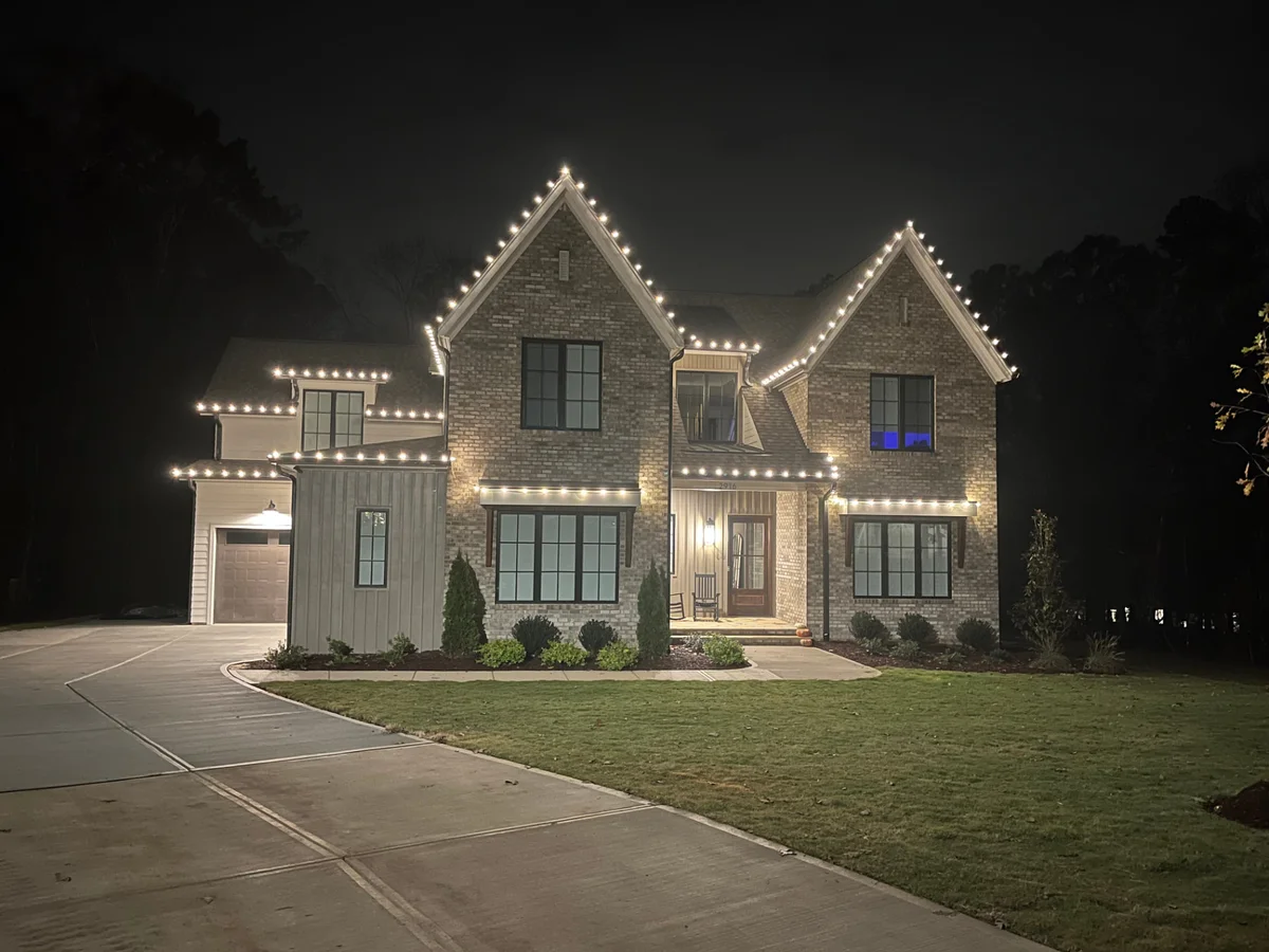 Warm white lights outlining every roofline and gable of a large two-story brick and board-and-batten home at night