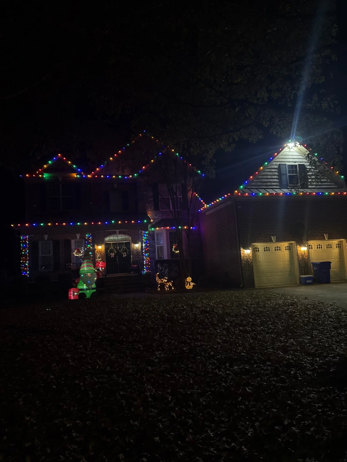 Multicolor lights along the roof peaks and wrapped porch columns of a brick two-story home with lit yard displays at night